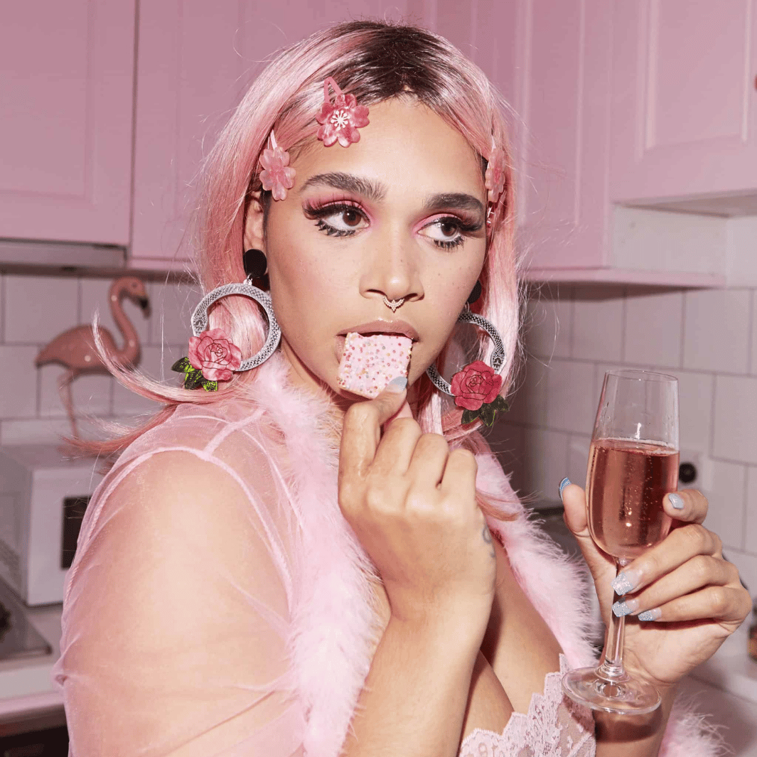 An image of a model wearing a pink sheer robe, pink straight hair and cherry blossom hair clips with silver Snake rose hoops and a glass of Rose. She is standing in a pink kitchen and eating a pink biscuit with 100s and 1000s. 