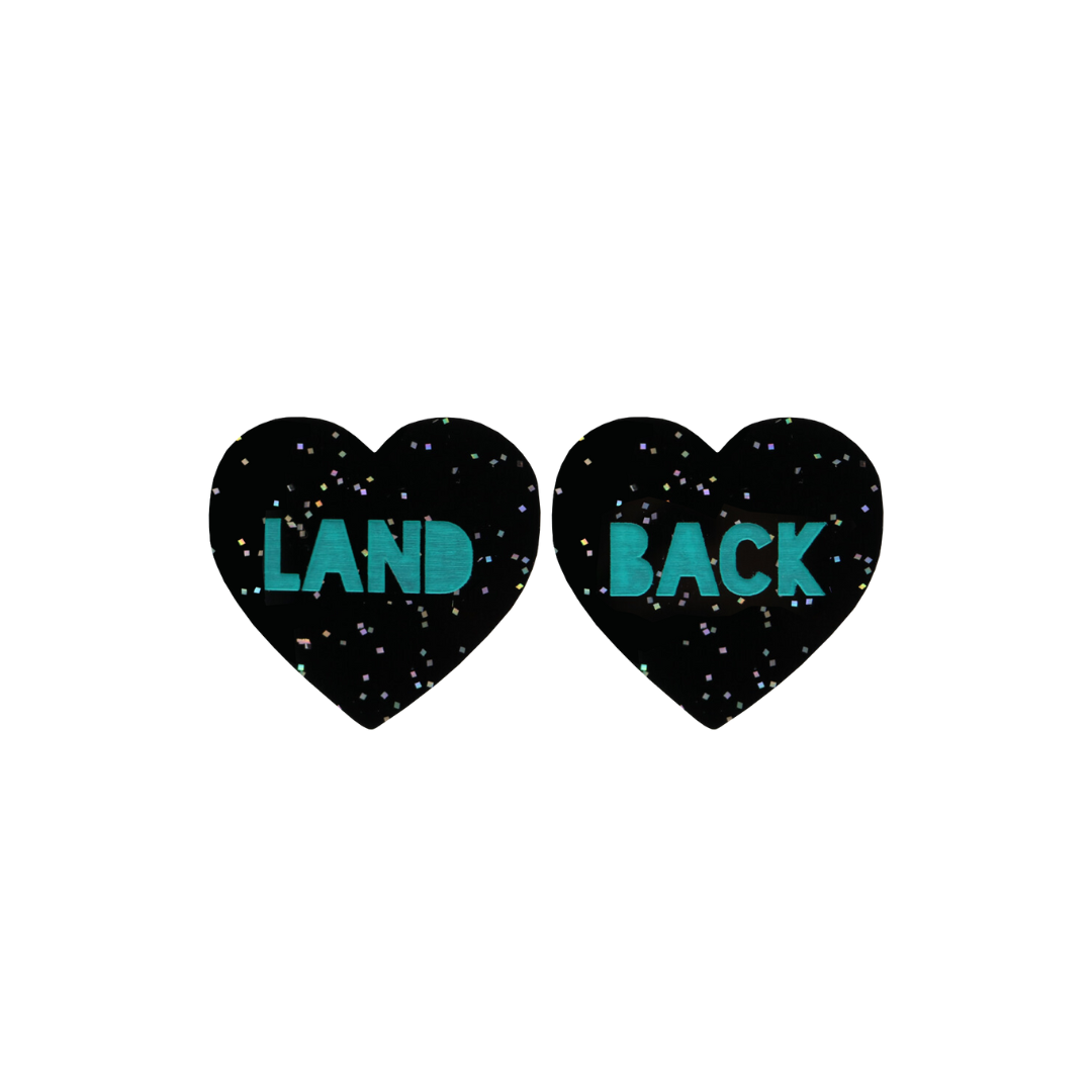 An image of Haus of Dizzy's Small ‘Land Back’ heart shaped dangle earrings, with Emerald Green “Land Back” text on a Black Glitter acrylic stud heart.