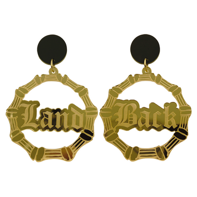 An image of Haus of Dizzy's large dangle bamboo hoop earrings, with Land back text on gold mirror acrylic and a matte black circle top.