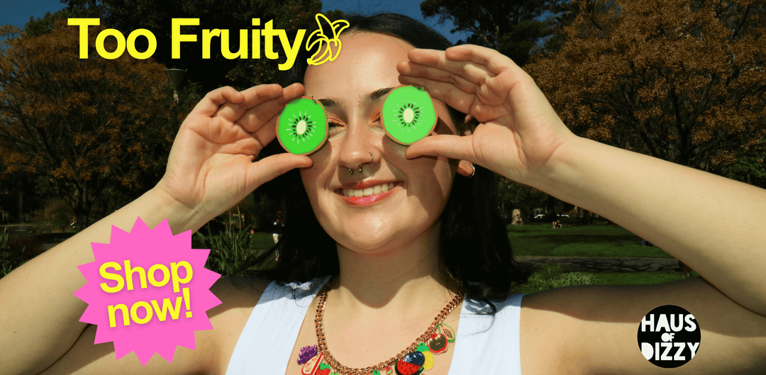 Smiling woman holds bright green kiwi slices over her eyes like glasses outdoors; colorful ad reads “Too Fruity,” “Shop now!” and Haus Dizzy.