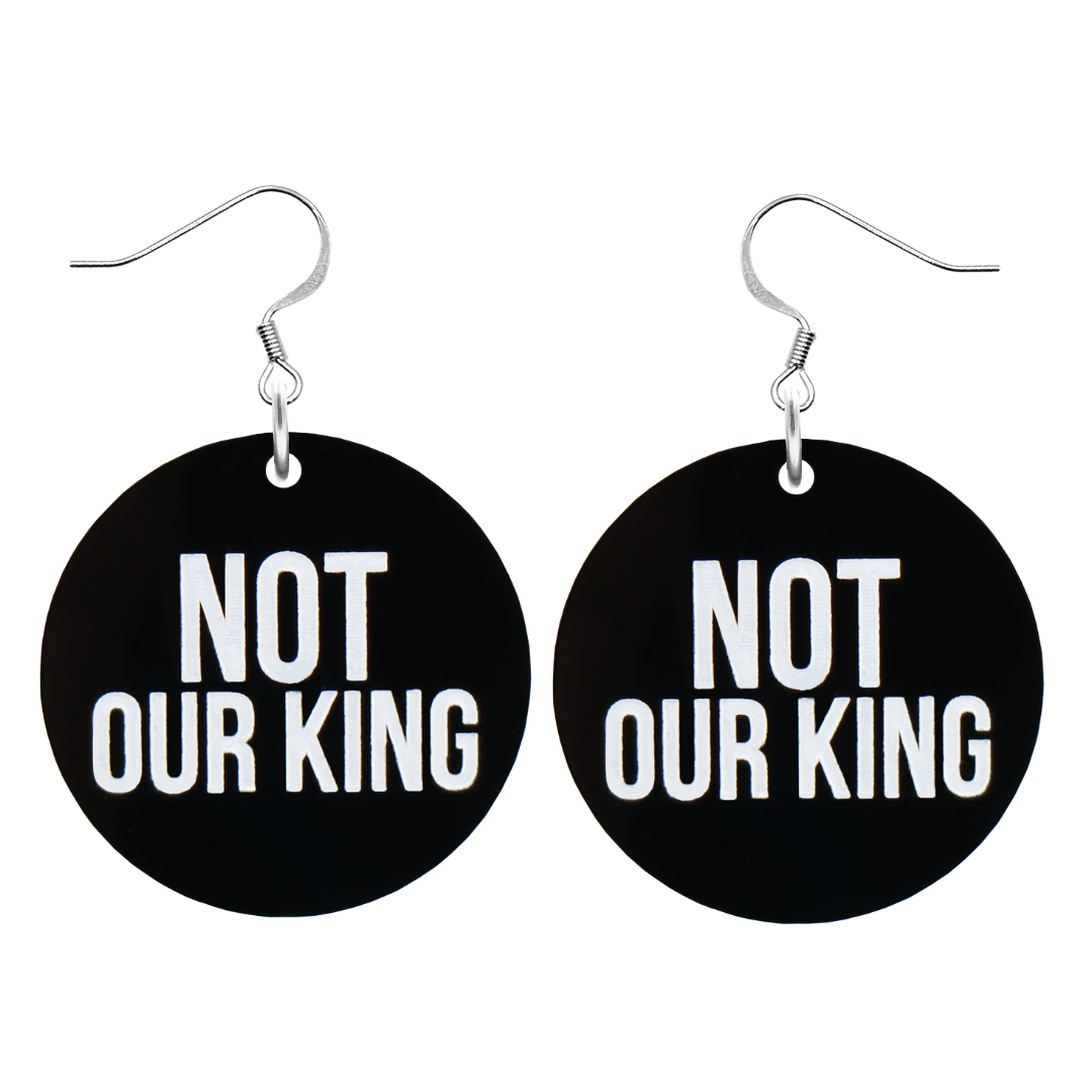An image of Haus of Dizzy's ‘Not our King’ circle shaped dangle earrings, with white paint fill Not our King text on black acrylic and a silver hook top.