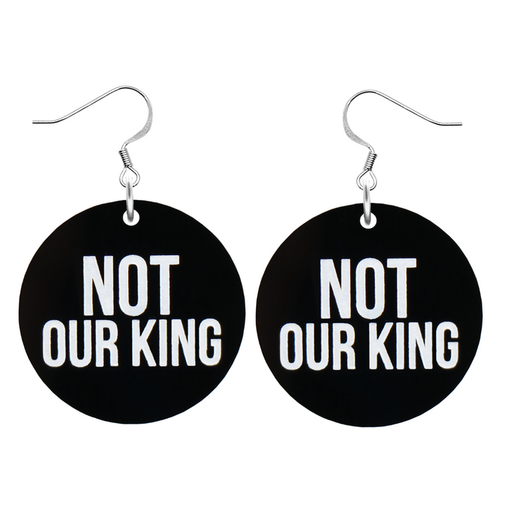 An image of Haus of Dizzy's ‘Not our King’ circle shaped dangle earrings, with white paint fill Not our King text on black acrylic and a silver hook top.