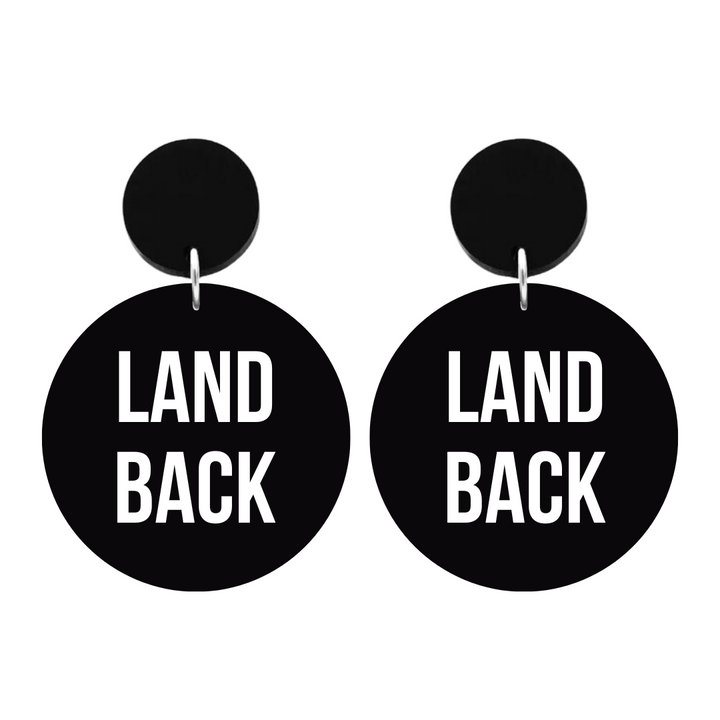 An image of Haus of Dizzy's ‘Land Back’ circle shaped dangle earrings, with white paint fill Land Back text on black acrylic and a black circle top.