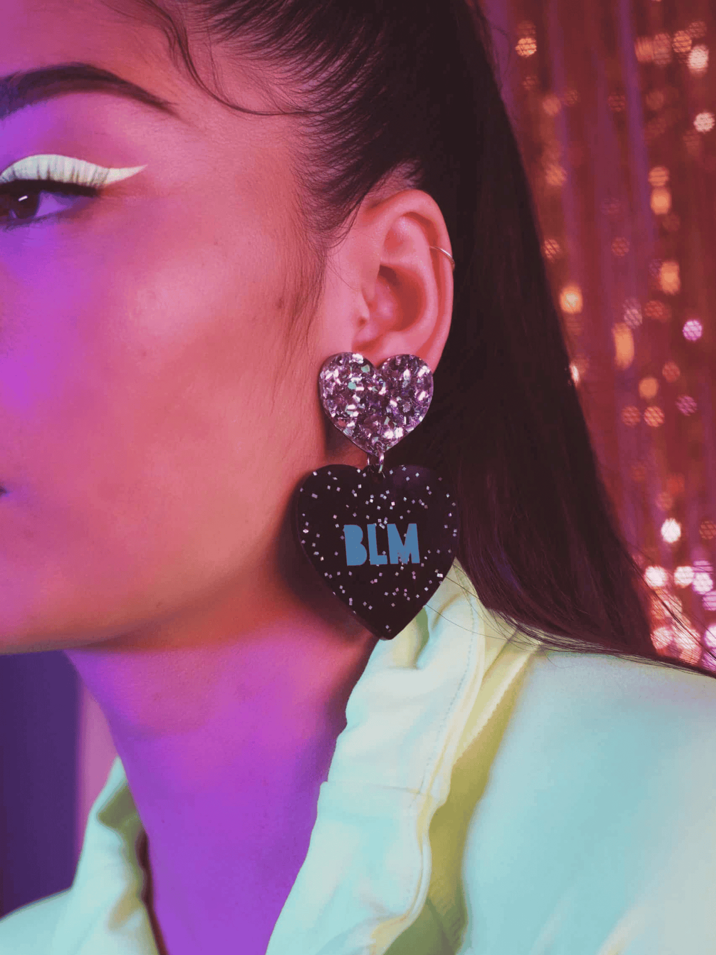 Model in neon yellow jacket wearing Haus of Dizzy's large 'BLM' dangle heart earrings with teal paint fill text etched into black glitter acrylic with a chunky silver glitter heart top.. 