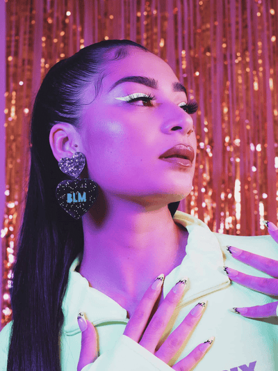 Model in neon yellow jacket and a high pony tail wearing Haus of Dizzy's large 'BLM' dangle heart earrings with teal paint fill text etched into black glitter acrylic with a chunky silver glitter heart top. Pictured in front of a gold and pink tinsel background.