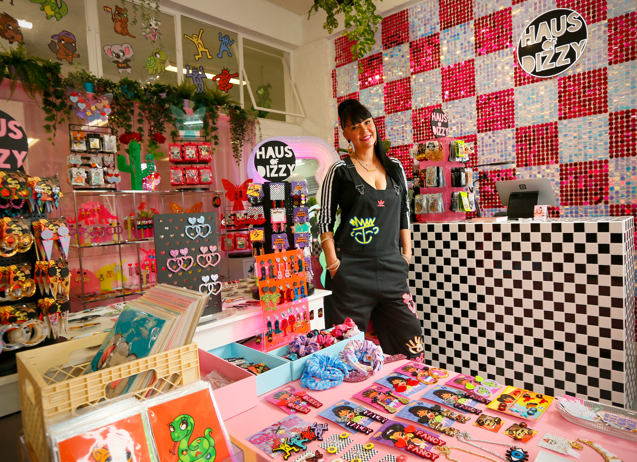 A photo of proud Wiradjuri woman, Kristy Dickinson standing in her colourful shop in front of her earring stands. The shop has a pink and white shimmer wall and earring displays. kristy is wearing a black Adidas Jumpsuit with colourful graffiti across it. her hair is black and tied in a high ponytail. 