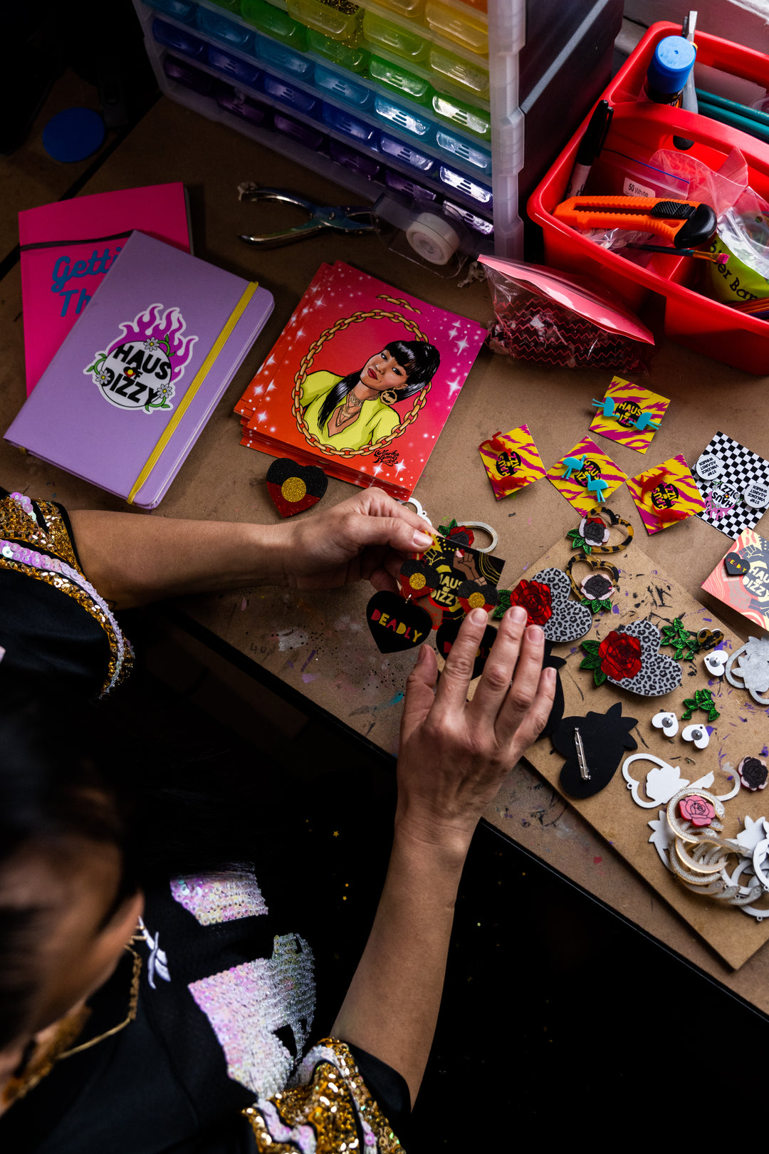 A photo of a proud Wiradjuri woman in her jewellery making studio. The image is focused on her hands making black heart acrylic earrings that say "Deadly" with an Aboriginal Flag circle stud top. Other earrings and pins are placed across the desk she is working on.