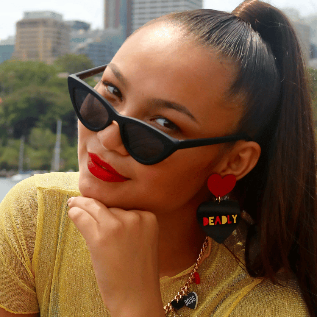A woman wearing red lipstick in a gold top with long brown hair tied in a high ponytail, wearing sunglasses and Haus of Dizzy's large Deadly dangle earrings with red glitter heart top, and red and yellow Deadly text. 