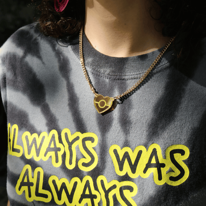 close up image of haus of dizzy mini mirror aboriginal flag pendant necklace on a model, wearing a grey tie dye t shirt. the necklace is reflecting in the sun