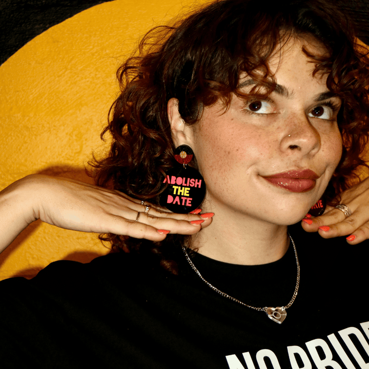 model wearing haus of dizzy black earring with 'Abolish The Date' text in aboriginal flag colours looking away from camera