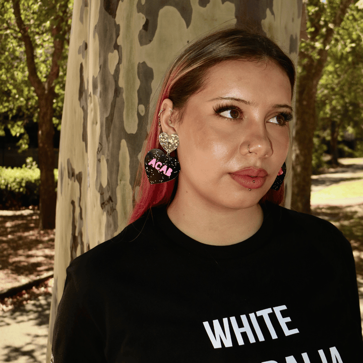 model wearing haus of dizzys acab glitter heart earrings with bold hot pink text and they are wearing a black t-shirt with white text, standing outdoors near trees.