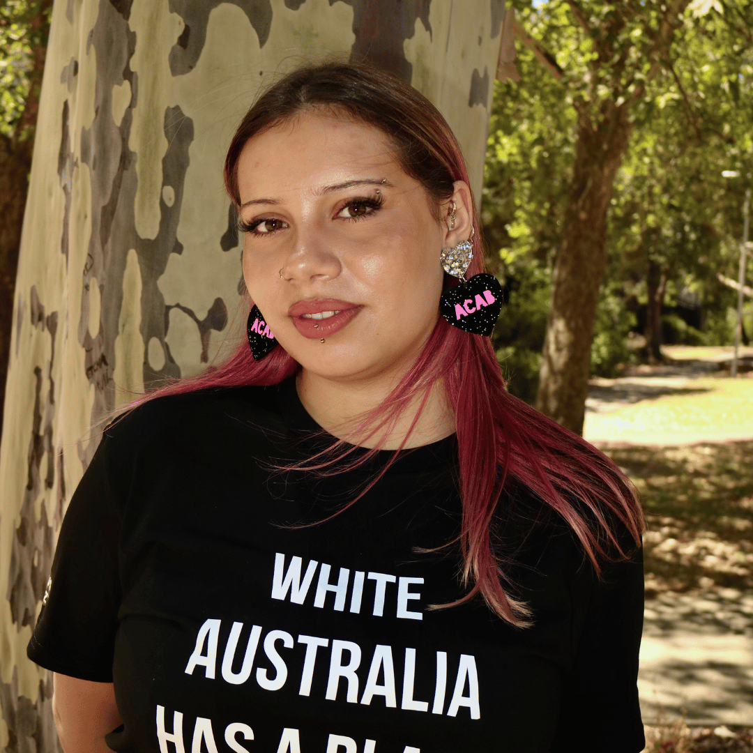 model wearing haus of dizzys acab glitter heart earrings with bold hot pink text and they are wearing a black t-shirt with white text, standing outdoors near trees.