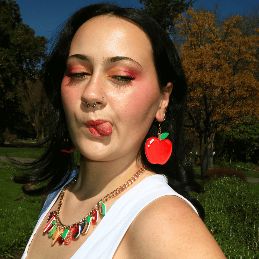 A woman outdoors wearing haus of dizzys an apple a day red apple-shaped dangling earring and a colorful candy-shaped pendant necklace, posing with her tongue sticking out against a background of green grass and autumn trees.