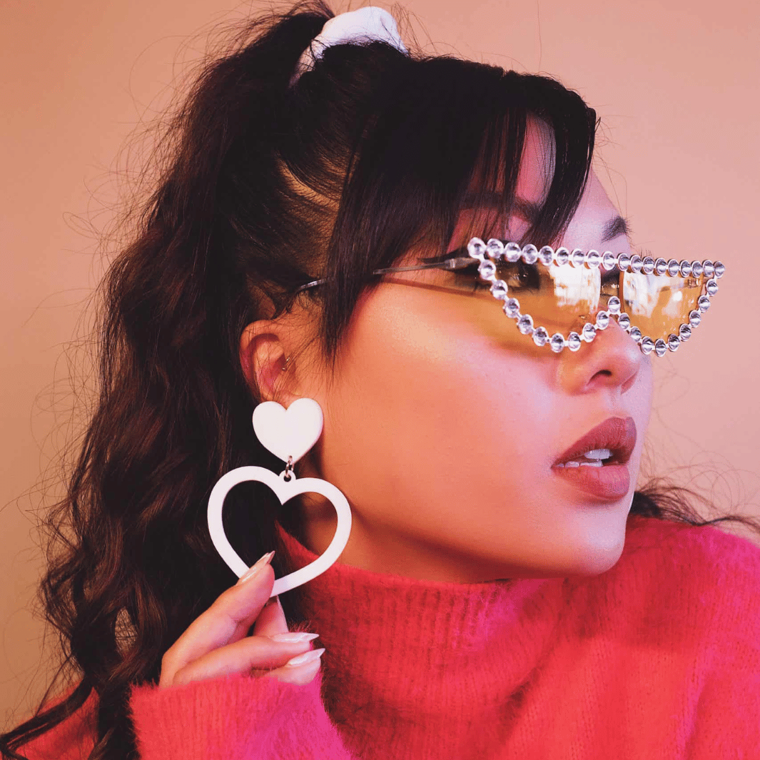 A model wearing a pink sweater and glasses, showcasing haus of dizzy white barbie heart earrings in size large