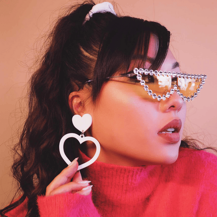 A model wearing a pink sweater and glasses, showcasing haus of dizzy white barbie heart earrings in size large