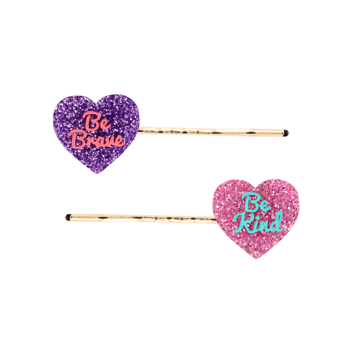 haus of dizzy heart shaped acrylic hair pins, one purple glitter with red 'be brave' text, the other pink glitter with aqua 'be kind' text, both on gold bobby hair pins