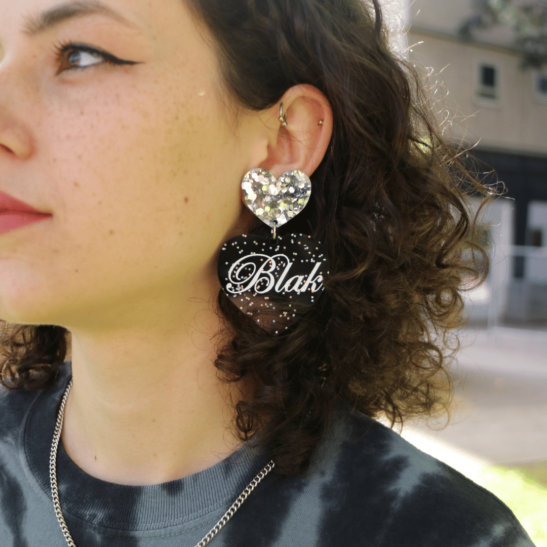close up of Aboriginal model wearing haus of dizzy blak glitter heart earrings with 'Blak' written in white paint script font