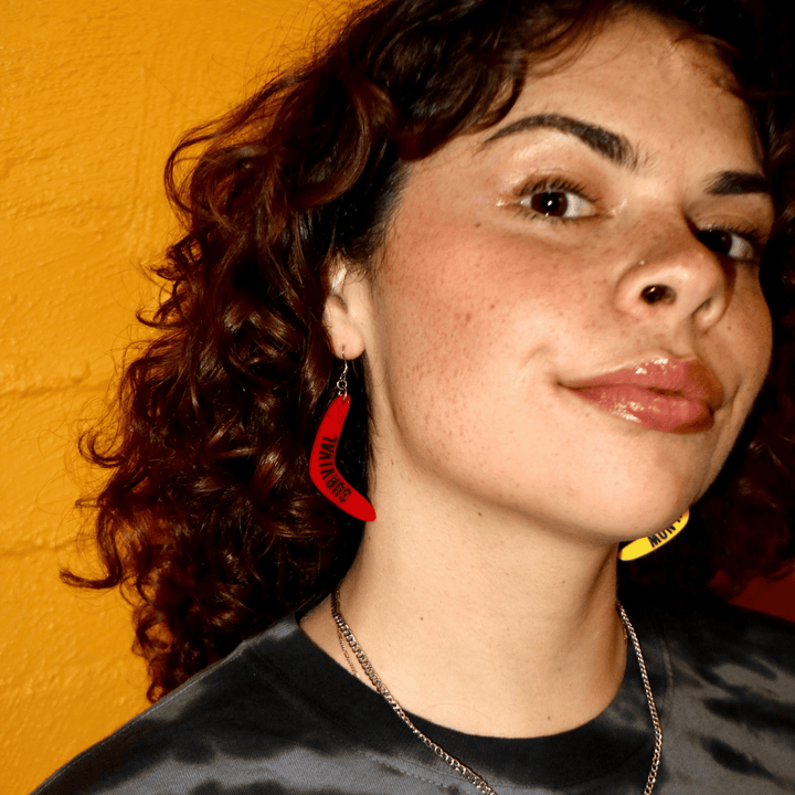 close up of model wearing haus of dizzy red boomerang earring that reads survival, this design is mob only