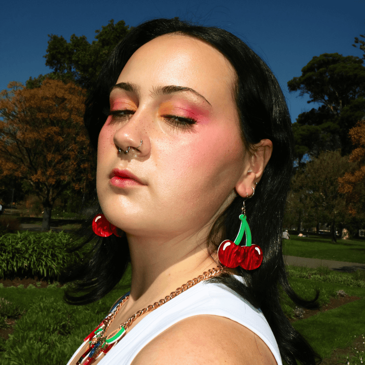 Close-up portrait of a person with black hair, bright pink makeup,  posing confidently in a sunny park wearing haus of dizzys cherry bomb cherry-shaped earrings 