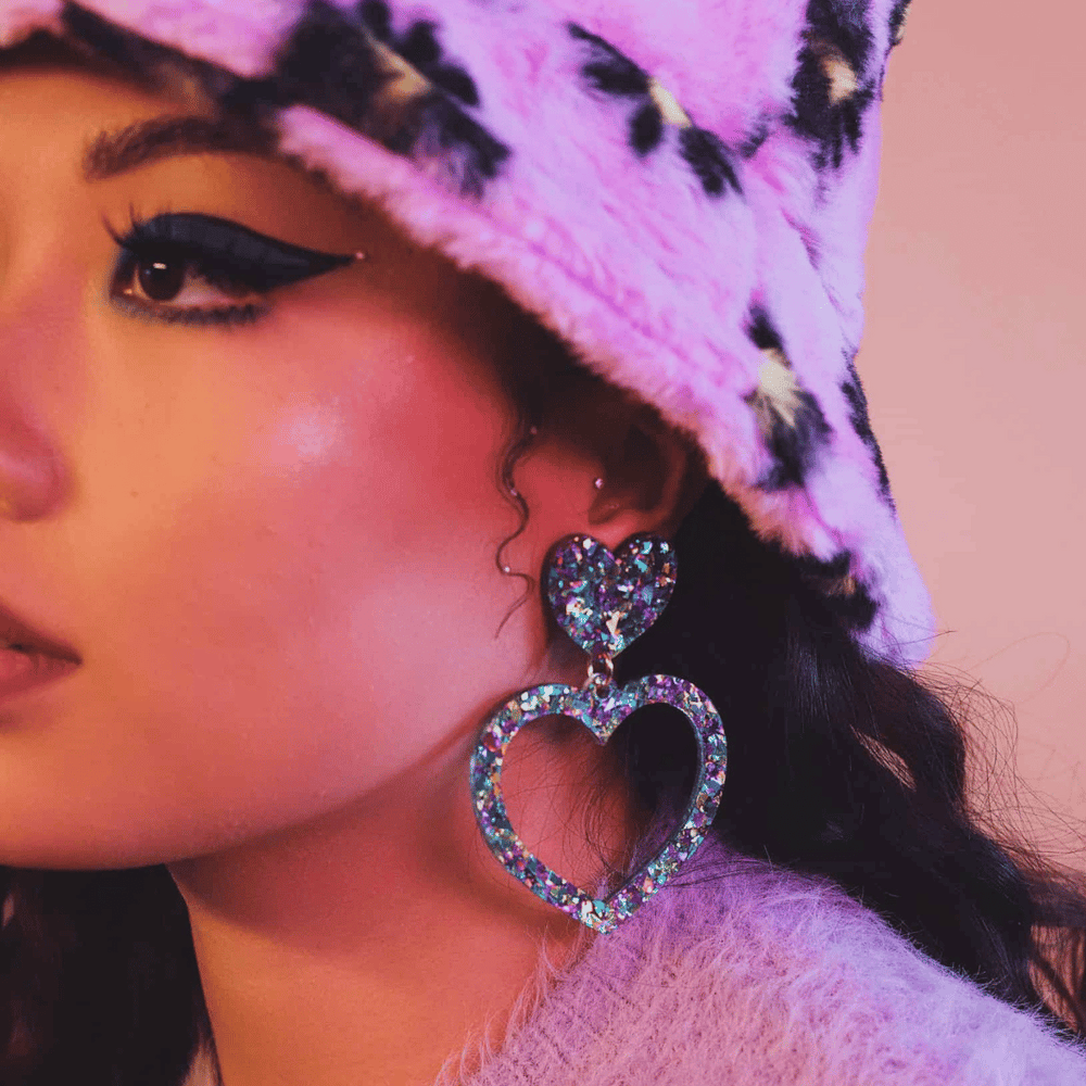 close up of model wearing haus of dizzy aquamarine crystal barbie heart earrings size large with fluffy printed hat