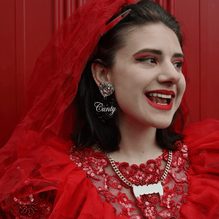 model in a red bride halloween costume wearing haus of dizzy 'cunty' black glitter heart earrings as well as haus of dizzy fang plate necklace halloween edition