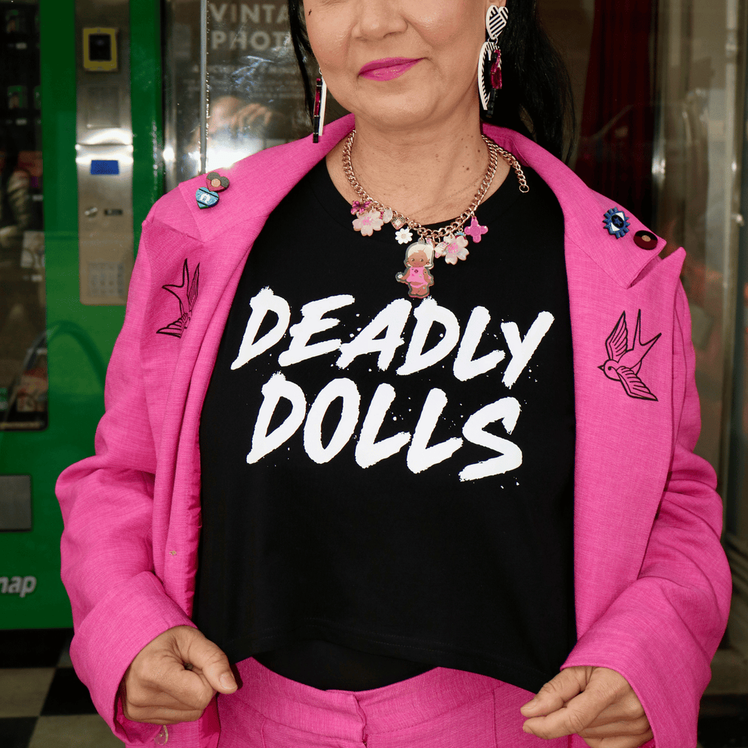 model wearing a bright pink tailored jacket and matching trousers over haus of dizzy's black crop t-shirt with large white text reading 'DEADLY DOLLS.' She is also wearing a haus of dizzy charm necklace and dangling flamingo heart earrings.