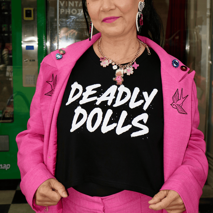 model wearing a bright pink tailored jacket and matching trousers over haus of dizzy's black crop t-shirt with large white text reading 'DEADLY DOLLS.' She is also wearing a haus of dizzy charm necklace and dangling flamingo heart earrings.