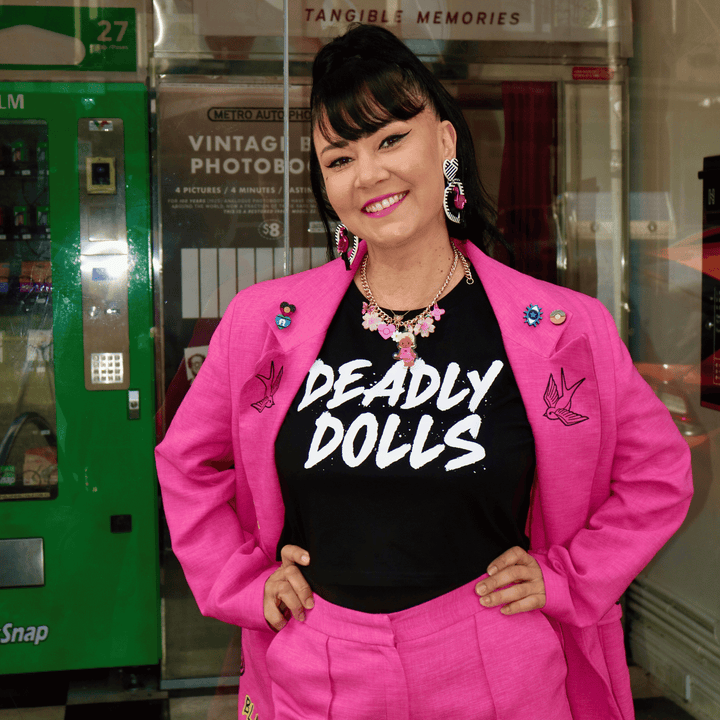 kristy of haus of dizzy posing in a bright pink blazer and matching pant set, wearing haus of dizzy's black crop tee that reads 'DEADLY DOLLS' in white paint brush font. She is standing in front of a glass storefront with a green camera vending machine visible on the left