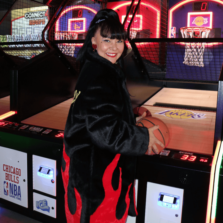 image of side of kristy from haus of dizzy wearing haus of dizzy x tibbs and bones deadly flamez fur jacket. it is a black knee length faux fur coat with red flames at the bottom she is looking at the camera while holding a basketball in an arcade