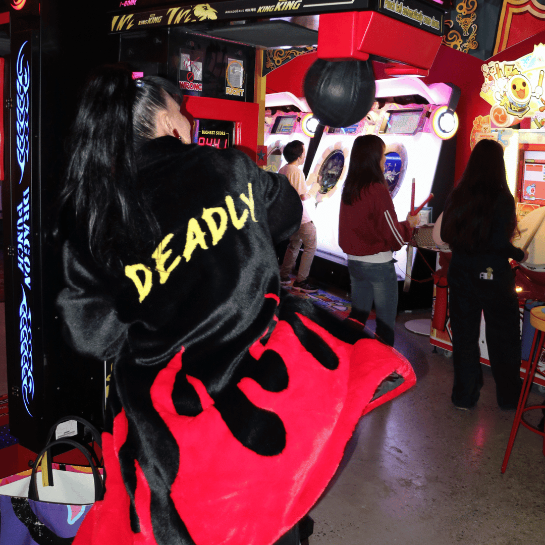 image of kristy from haus of dizzy wearing haus of dizzy x tibbs and bones deadly flamez fur jacket. she is punching a boxing game in the arcade. the coat is a black knee length faux fur coat with red flames at the bottom and on the back it has 'deadly' in graffiti style yellow font