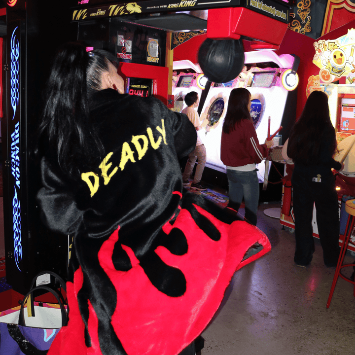 image of kristy from haus of dizzy wearing haus of dizzy x tibbs and bones deadly flamez fur jacket. she is punching a boxing game in the arcade. the coat is a black knee length faux fur coat with red flames at the bottom and on the back it has 'deadly' in graffiti style yellow font