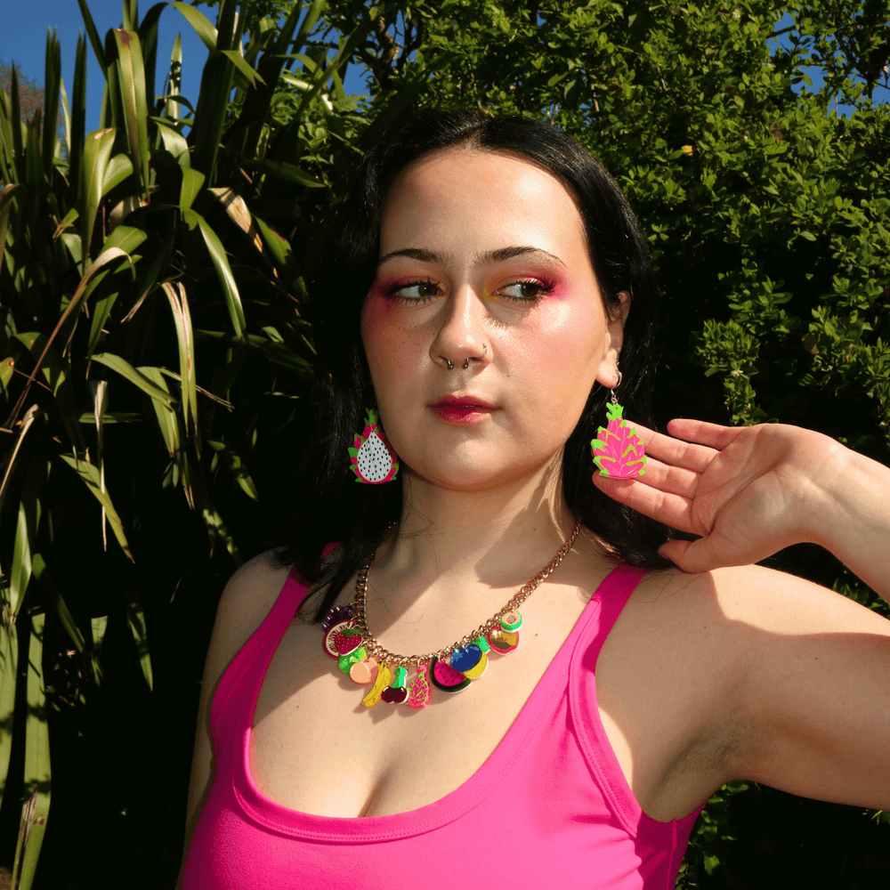 chest up shot of model wearing haus of dizzy's dragonfruit dangle earrings in front of a bright green bush wearing a hot pink tank top and fruit charm necklace.