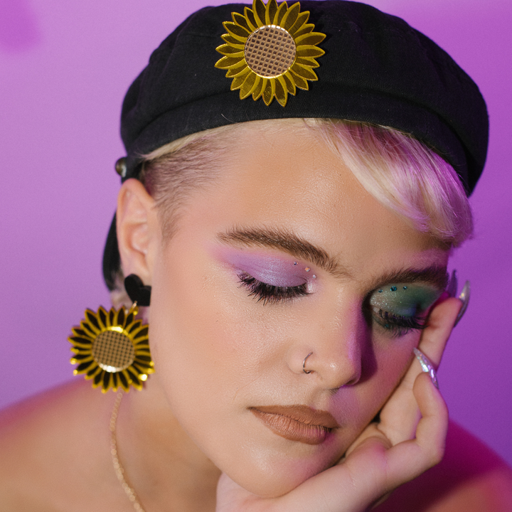 A model wearing haus of dizzys yellow mirrored sunflower earrings with a black hat and makeup, with the earrings visible in the foreground.