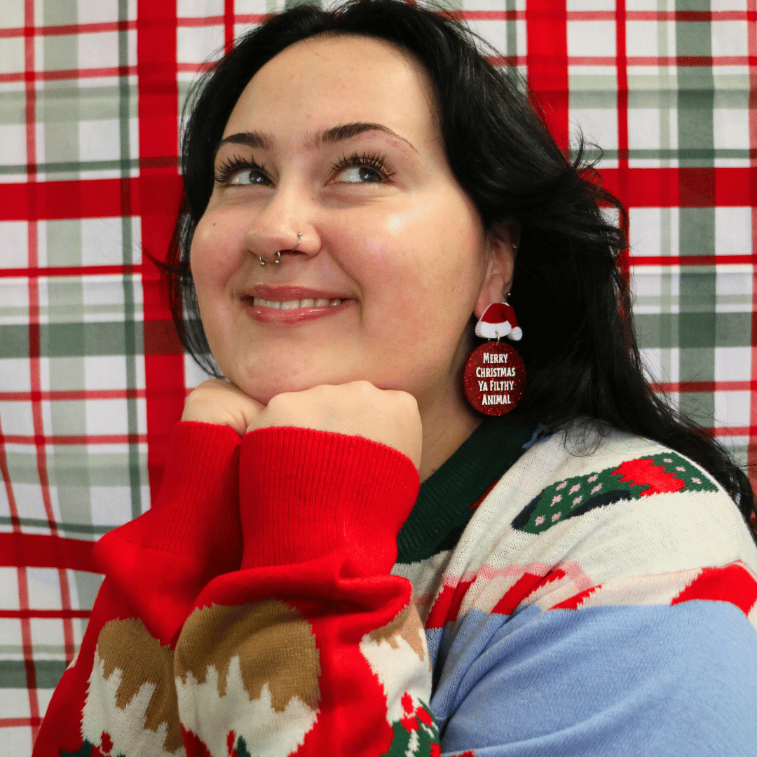 model wearing ugly christmas sweater in front of plaid red green background. she is wearing haus of dizzy filthy animal glitter earrings with santa hats looking away from camera