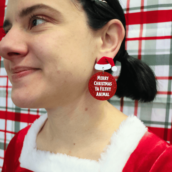 model wearing santa outfit in front of plaid red green background. she is wearing haus of dizzy filthy animal glitter earrings with santa hats looking away from camera