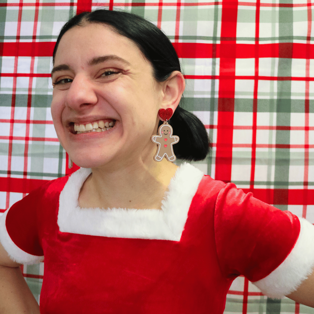 haus of dizzy model wearing santa outfit against a red green plaid background, wearing haus of dizzy gingerbread friend earrings, grinning at the camera