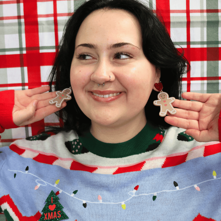 haus of dizzy model wearing ugly christmas sweater against a red green plaid background, wearing haus of dizzy gingerbread friend earrings, smiling looking away from the camera and holding the earrings to face the camera