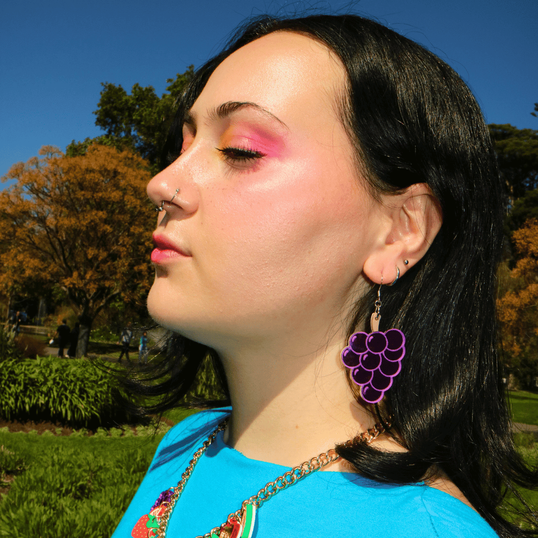 close up side profile shot of model wearing haus of dizzy's pre wine, grape shaped dangle earrings which are made out of purple mirror acrylic and matte brown acrylic for the stem. the model has bright pink/yellow makeup and a bright blue top on.