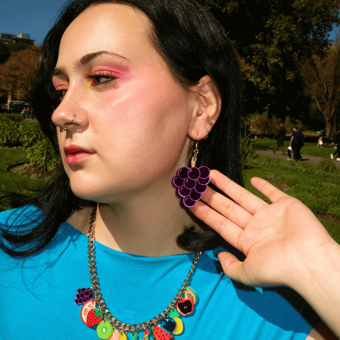 close up shot of model wearing haus of dizzy's pre wine, grape shaped dangle earrings which are made out of purple mirror acrylic and matte brown acrylic for the stem. the model is holding the earrings in place while looking away from the camera