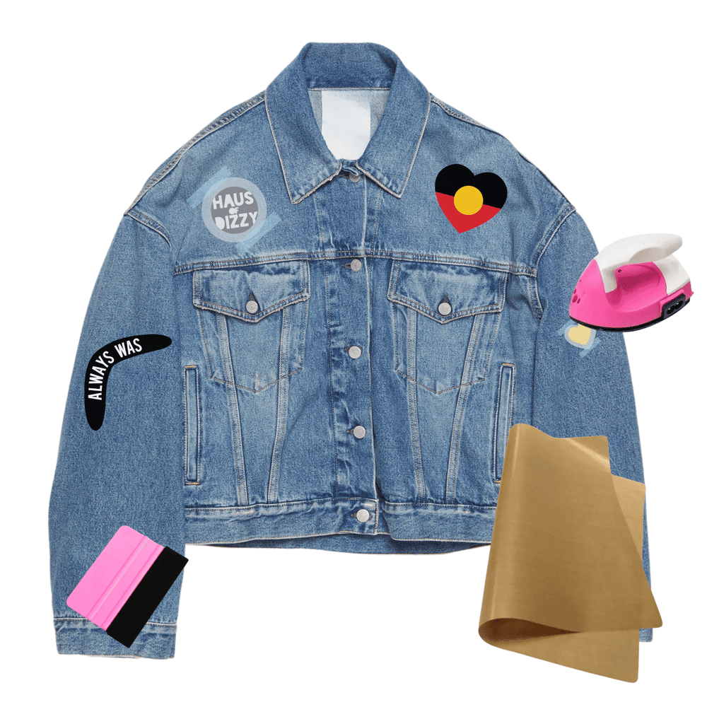 A light blue denim jacket with multiple colorful iron on prints/decals, displayed alongside props including a pink iron, brown teflon paper, and a plastic squeegee