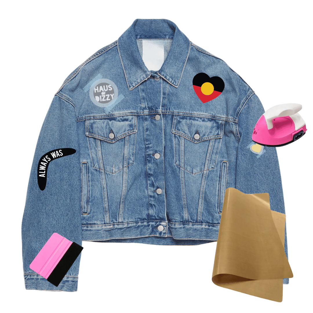 A light blue denim jacket with multiple colorful iron on prints/decals, displayed alongside props including a pink iron, brown teflon paper, and a plastic squeegee