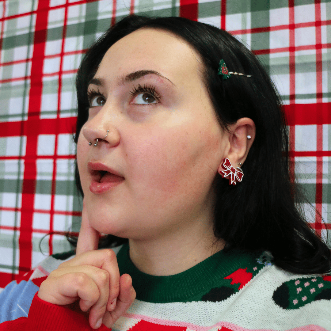 model wearing ugly christmas sweater with haus of dizzy jolly bow stud earrings in red glitter looking away from camera