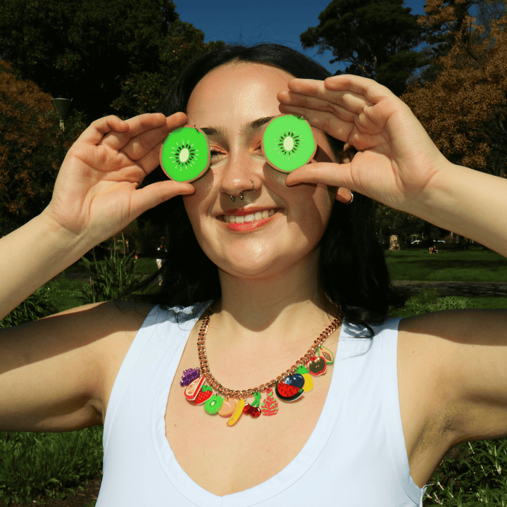 A smiling woman outdoors wearing a colorful fruit-themed charm necklace, holding haus of dizzy kiwi slice earrings over her eyes in a playful pose against a sunny park background.