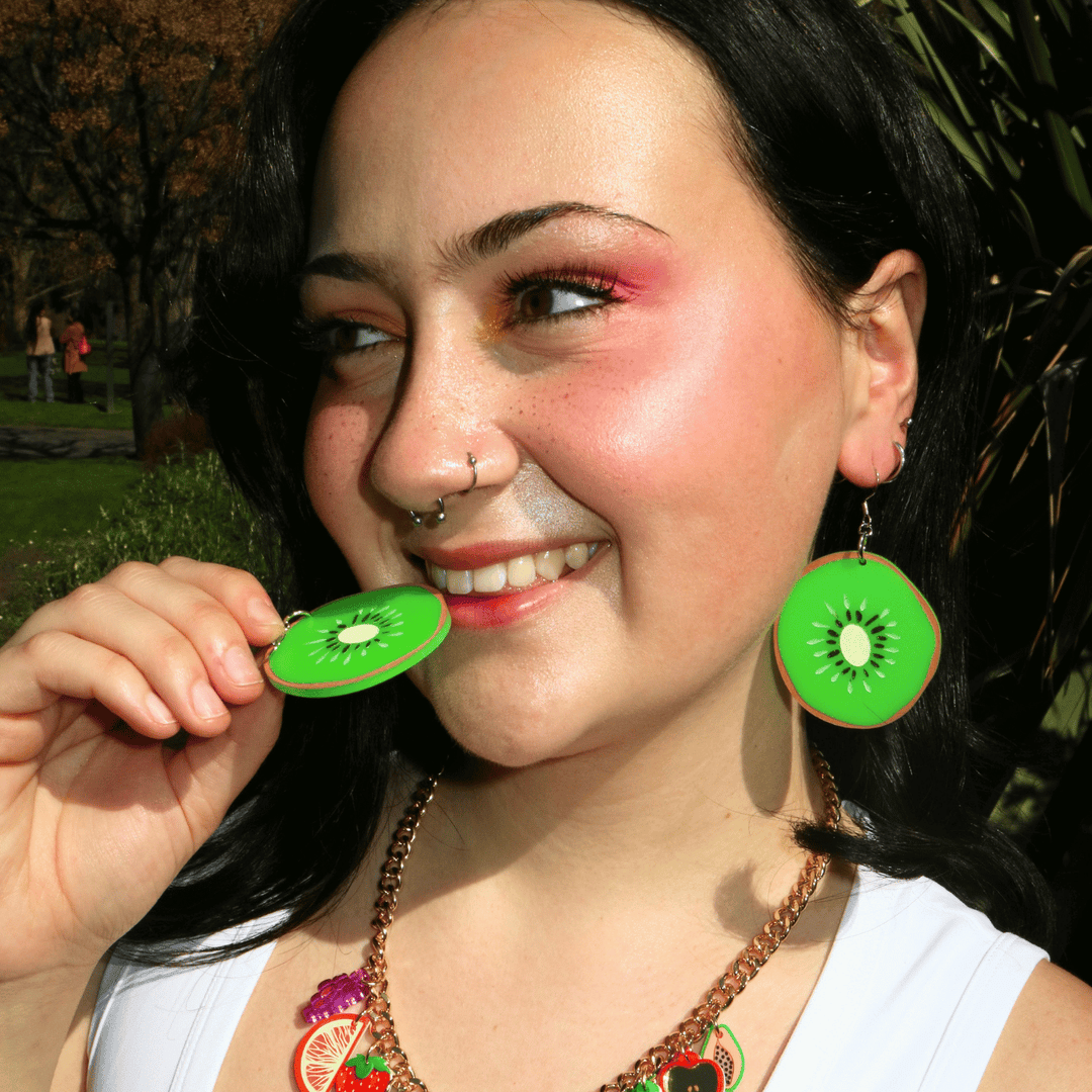 close up shot of model looking away from camera wearing haus of dizzy;s kiwi shaped dangle earrings, with one in her ear the other pretending to take a bite of. the earrings are made of lime green matte acrylic with hand painted brown black and ivory details.