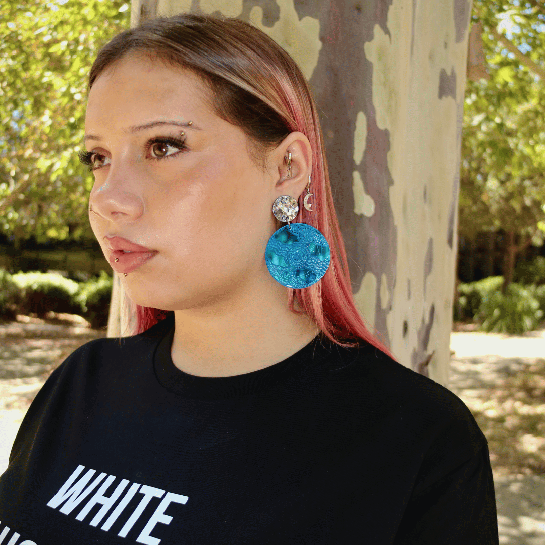 model wearing haus of dizzy x kiya watt collaboration earrings in blue mirror with aboriginal artwork engraved into blue mirror acrylic with chunky silver glitter circle stud top. 