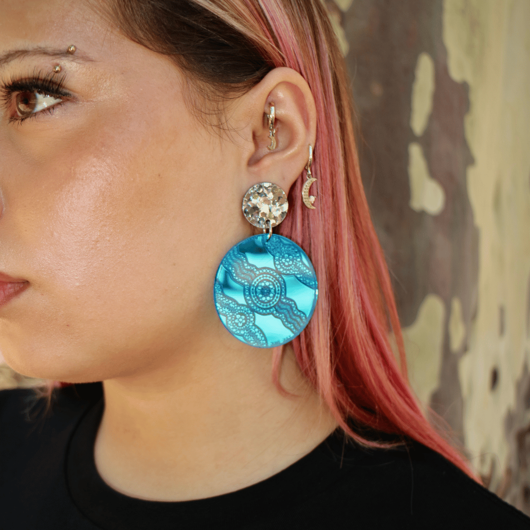 close up image of model wearing haus of dizzy x kiya watt collaboration earrings in blue mirror with aboriginal artwork engraved into blue mirror acrylic with chunky silver glitter circle stud top. 