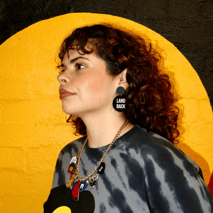 an image of a model from the shoulders up wearing haus of dizzys 'land back' earrings as part of the wear the resistance collections. they are plain black circle earrings with bold white text. they are for Mob only.