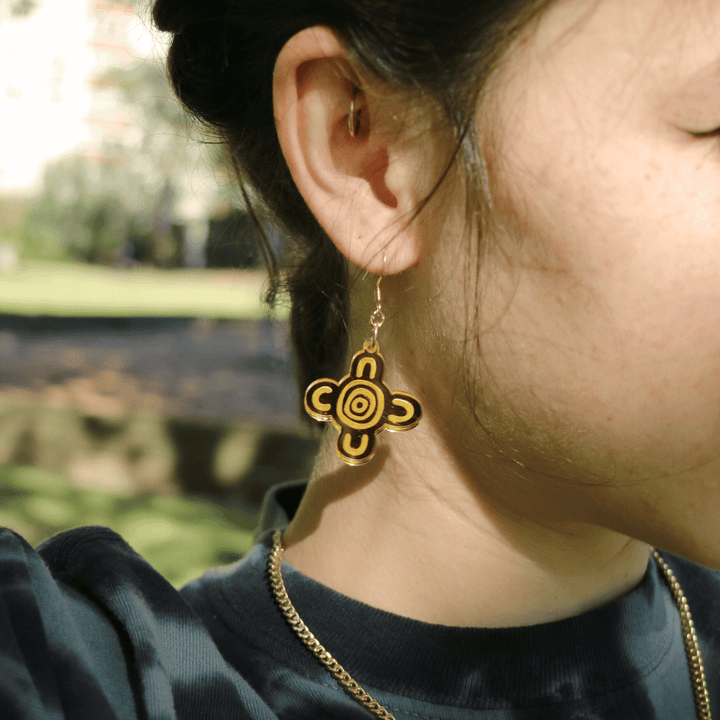 close up of model wearing haus of dizzy meeting place mirror earrings size small  in gold mirror with a blurred natural background. the earring features aboriginal art which represents people coming together, seated at a specific site, camp or gathering place.