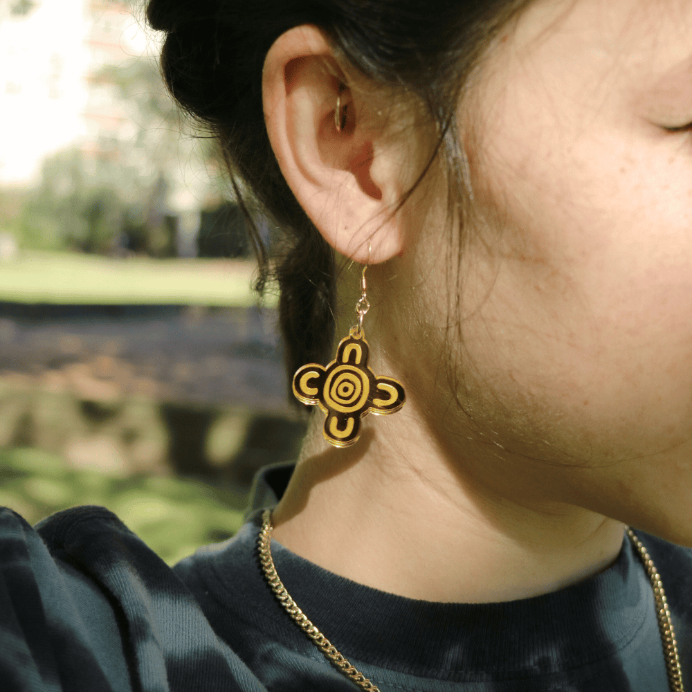 close up of model wearing haus of dizzy meeting place mirror earrings size small with a blurred natural background. the earring features aboriginal art which represents people coming together, seated at a specific site, camp or gathering place.