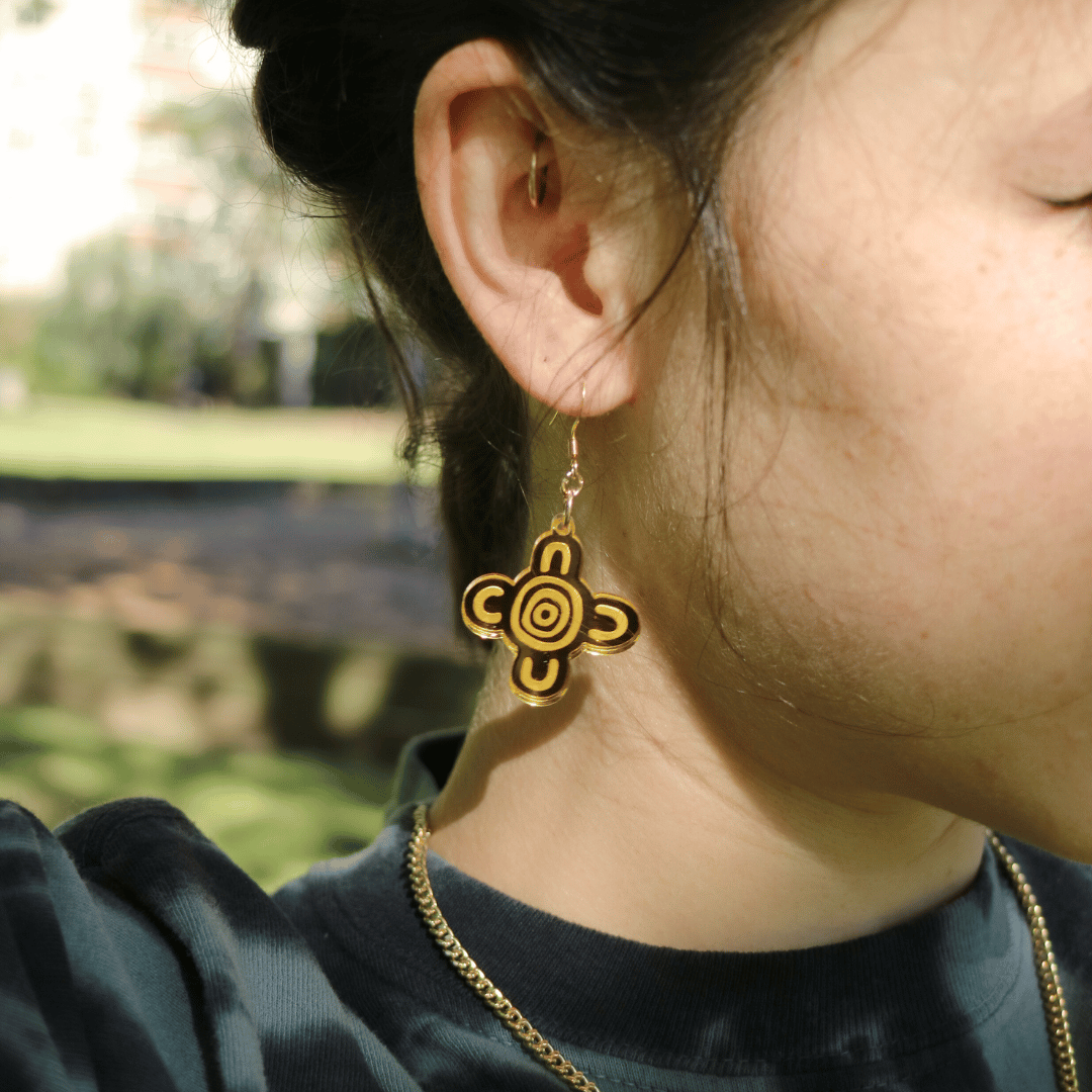 close up of model wearing haus of dizzy meeting place mirror earrings size small with a blurred natural background. the earring features aboriginal art which represents people coming together, seated at a specific site, camp or gathering place.